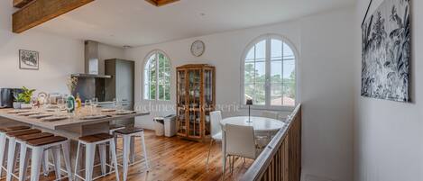 A spacious kitchen with wooden beams, a dining area with white chairs, and large windows offering views of greenery.