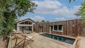 Una terraza de madera con piscina, un comedor y una casa con grandes ventanales.
