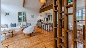 Amplia sala de estar con piso de madera, un sofá, una silla y un comedor con taburetes. Amplias ventanas ofrecen luz natural y vistas al exterior.
