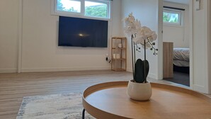 Ein modernes Zimmer mit großem Fenster, Flachbildfernseher, Holzboden und rundem Holztisch mit orchideenhaltiger Pflanze.