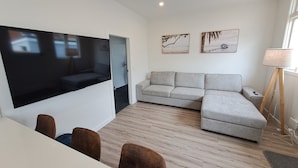 Ein modernes Wohnzimmer mit einem Flachbildfernseher, einem grauen Ecksofa, einem Esstisch mit Stühlen und einer Stehlampe.