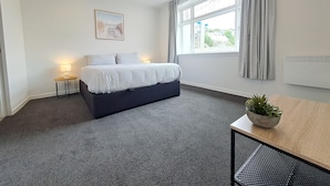 Ein modernes Schlafzimmer mit einem Bett, einem kleinen Tisch mit Pflanze, einem Nachttisch, einem Fenster mit Vorhängen und einem an der Wand hängenden Bild.