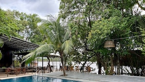 A poolside area with a covered seating area, tables, and chairs, surrounded by lush greenery and trees.