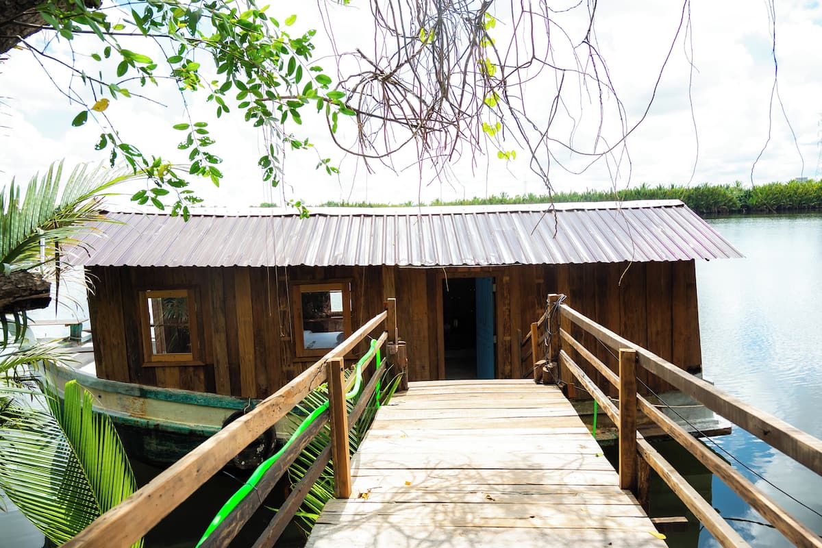 A wooden dock leading to a small wooden building with a metal roof, surrounded by water and greenery.