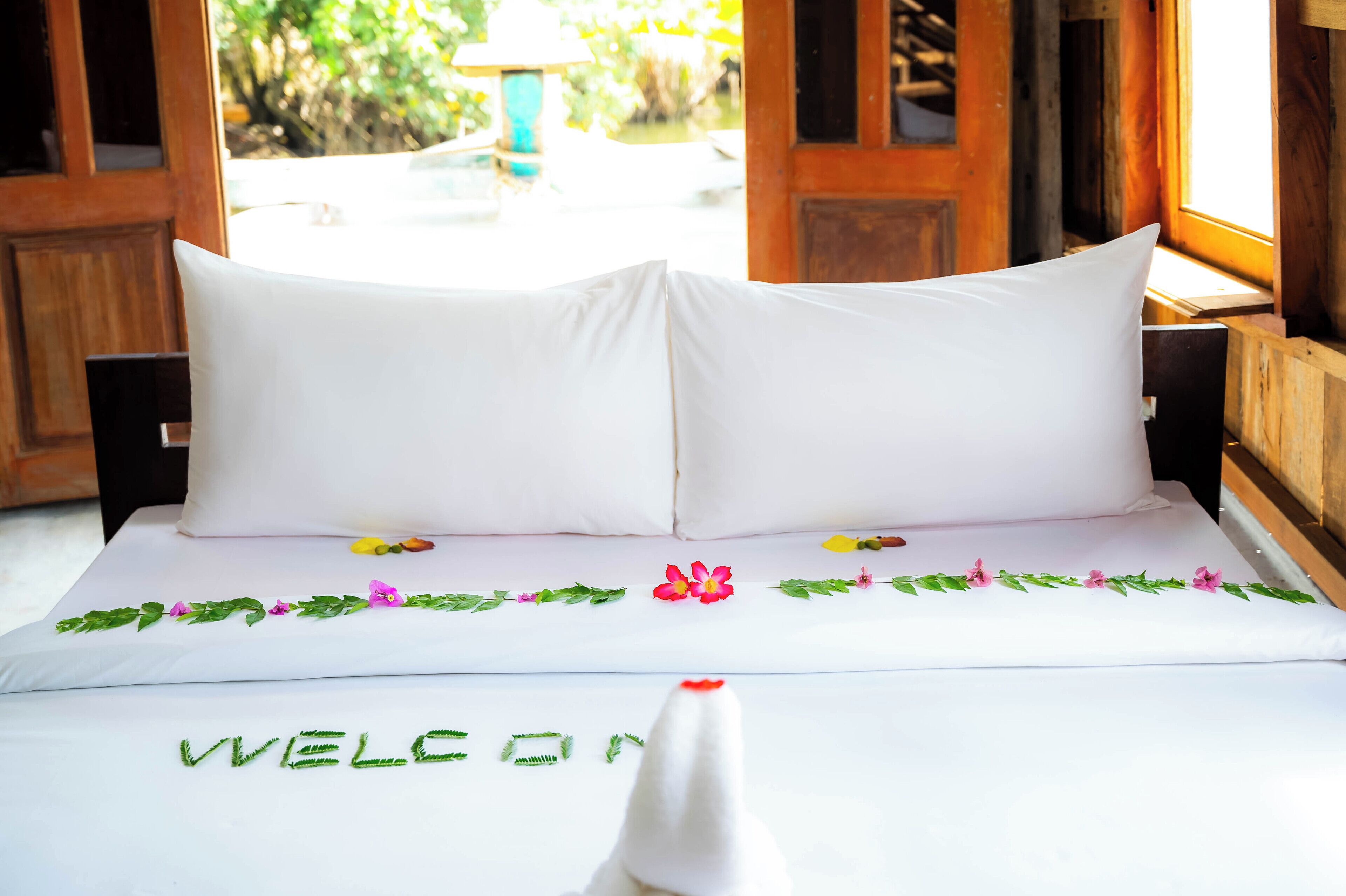 A bed with white linens and a "WELCOME" sign, wooden headboard, and floral decorations.
