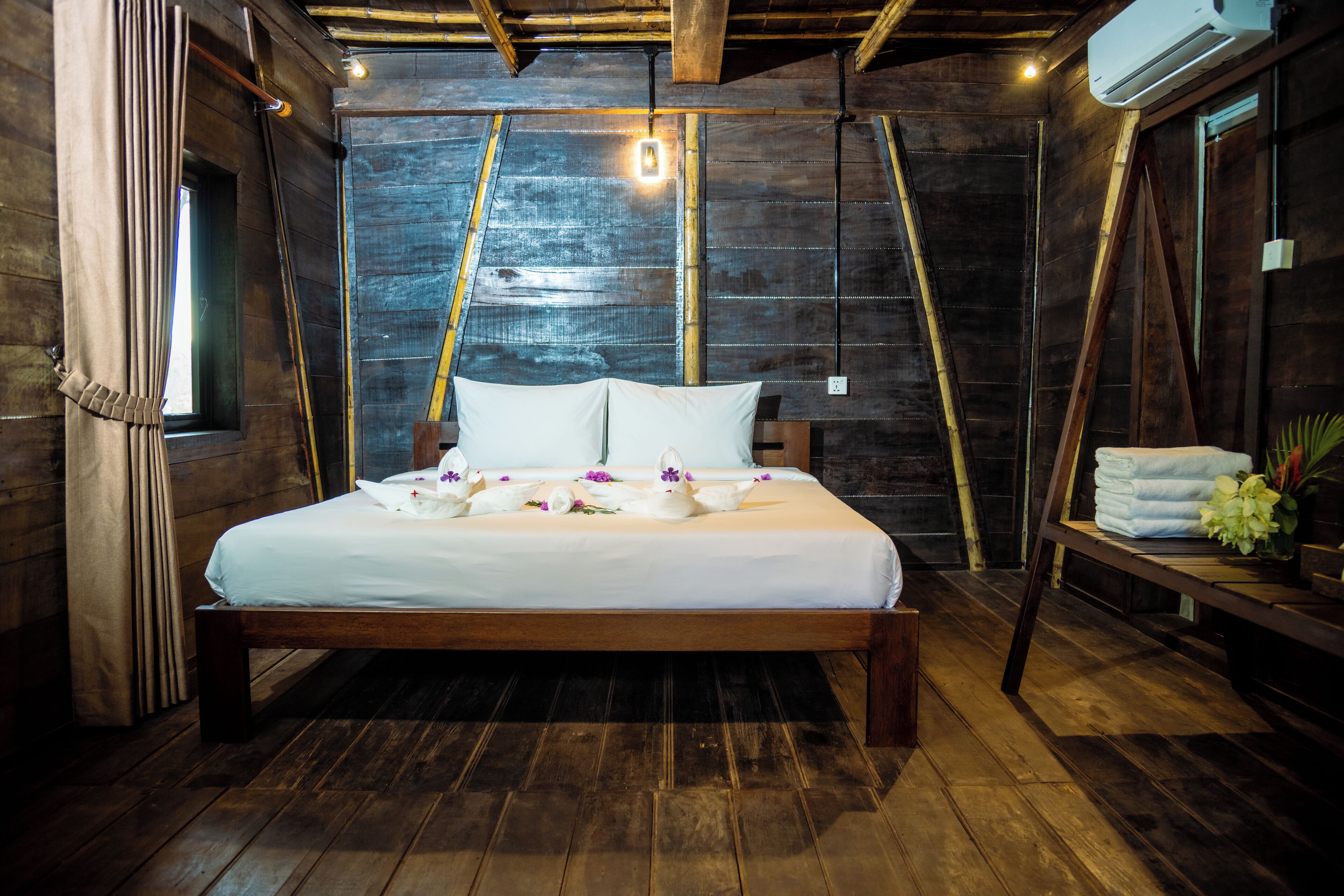 A wooden-framed bed with white linens and decorative swans, a wooden floor, and a rustic wall with a window and an air conditioning unit.