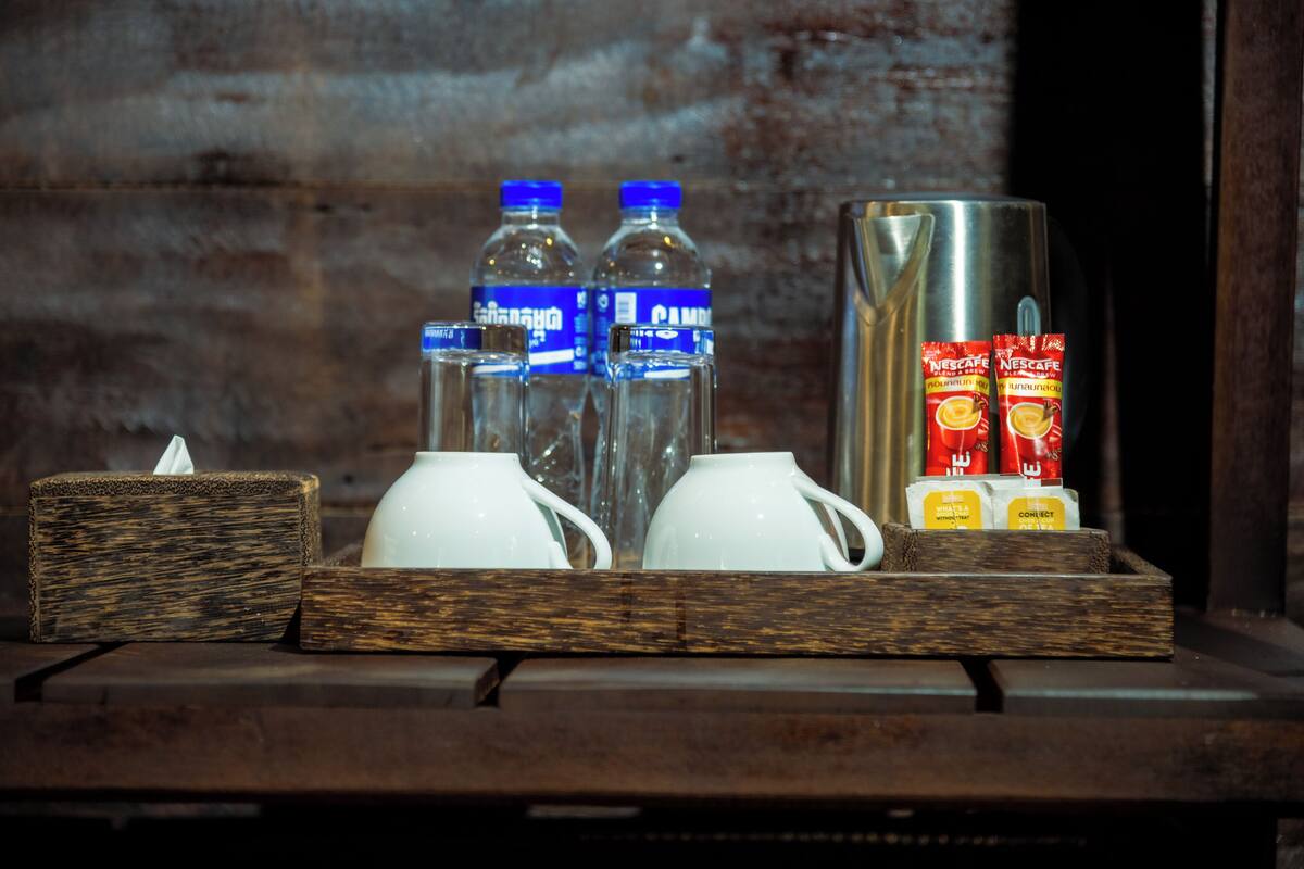 A wooden tray with a tissue box, two water bottles, two white cups, a stainless steel coffee pot, and two packets of Nescafe instant coffee.