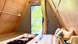 A cozy, triangular tent-style room with a neatly made bed, a small table, and a view of greenery outside.
