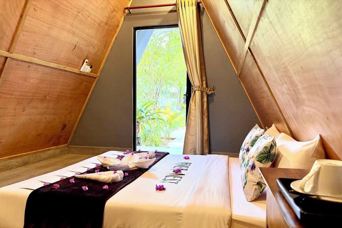 A cozy, triangular tent-style room with a neatly made bed, a small table, and a view of greenery outside.