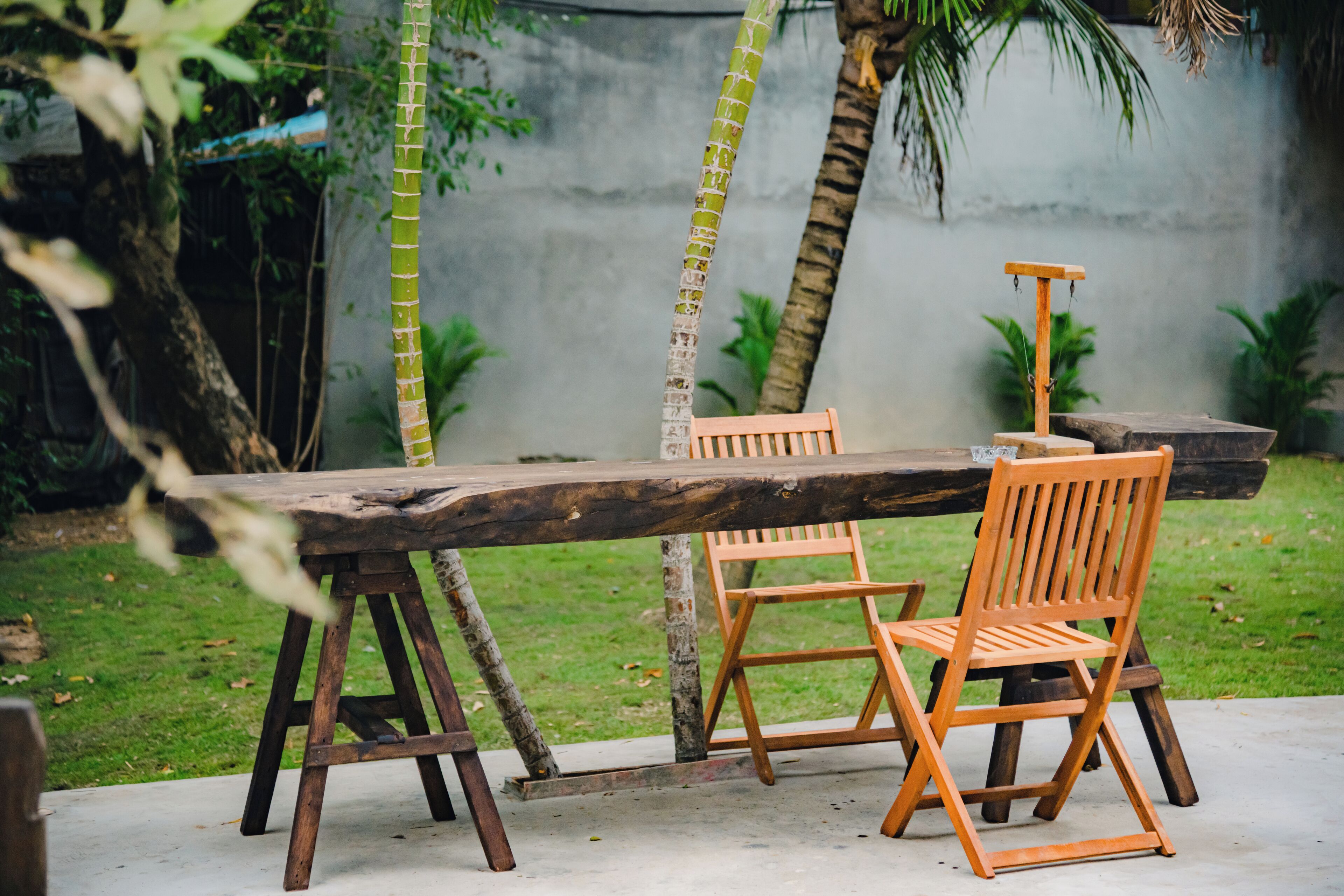 Un espace de restauration extérieur de style rustique, avec une table et des chaises en bois, entouré de verdure et bordé d’un mur en béton.