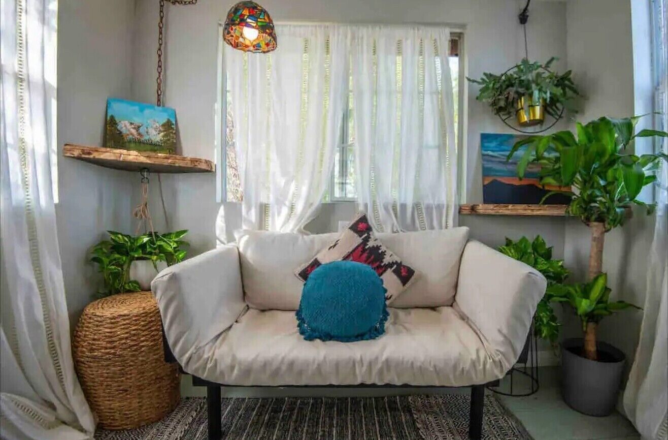 Een gezellige woonkamer met een witte bank, een blauw kussen en een hanglamp. Er staan planten en kunstwerken op planken, en er ligt een gevlochten mand op de vloer.