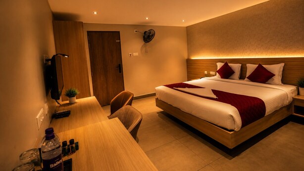 A hotel room with a large bed, a desk with a chair, a TV, and a wall-mounted fan. - Hypolux Business Hotel (Palakkad)
