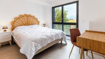 Une chambre avec un grand lit, une tête de lit en bois, une table de chevet avec une lampe, une table en bois et une fenêtre donnant sur des arbres.