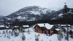 A log cabin with a snow-covered roof, surrounded by a forest and mountains in the background.