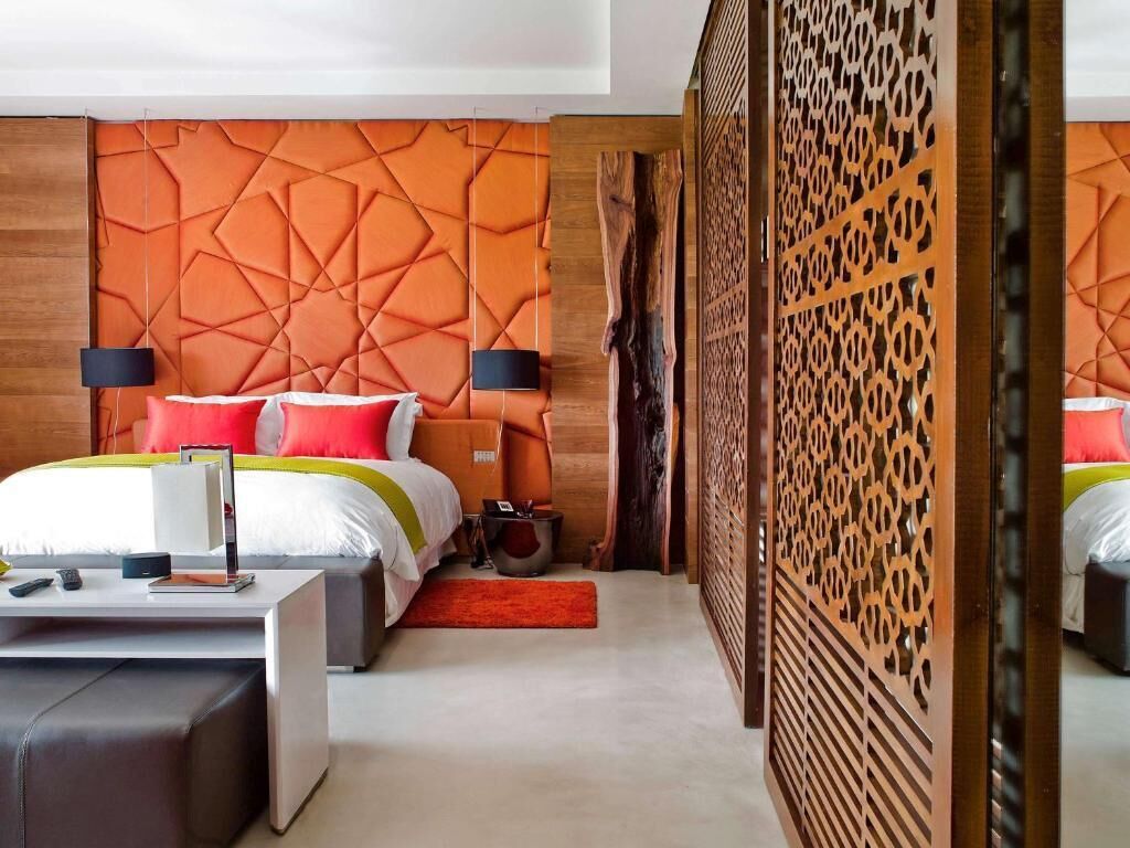 Une chambre d’hôtel moderne avec une tête de lit à motifs géométriques, un grand lit avec une literie rouge et blanche, une table de chevet avec une lampe et une cloison en bois ornée de motifs complexes découpés.