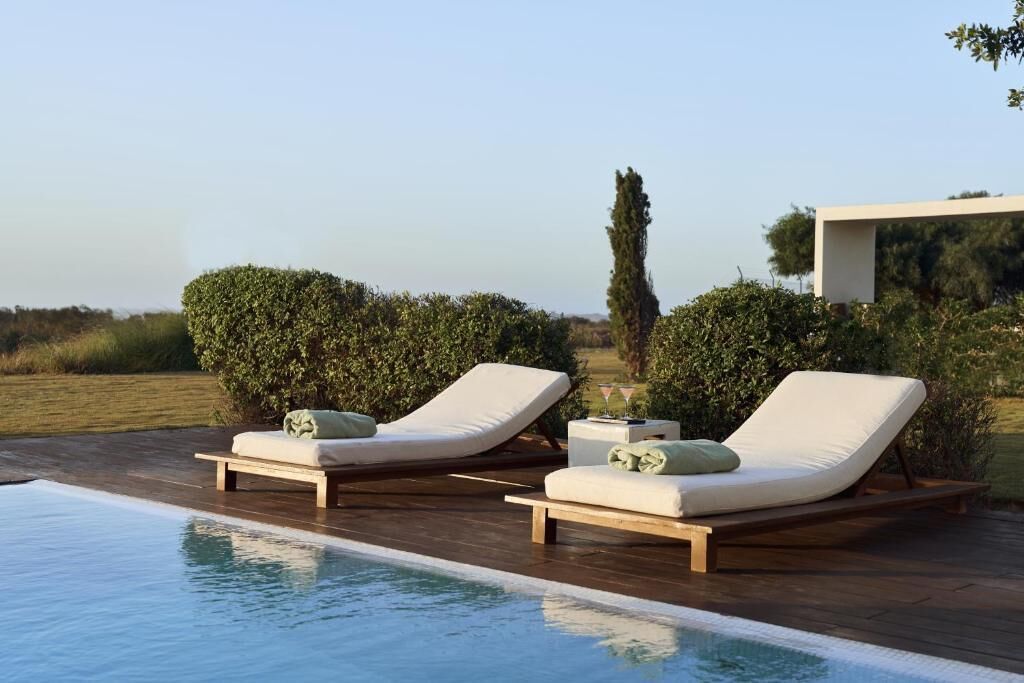 Area piscina con due lettini e un tavolino, circondata da vegetazione e un cielo sereno.