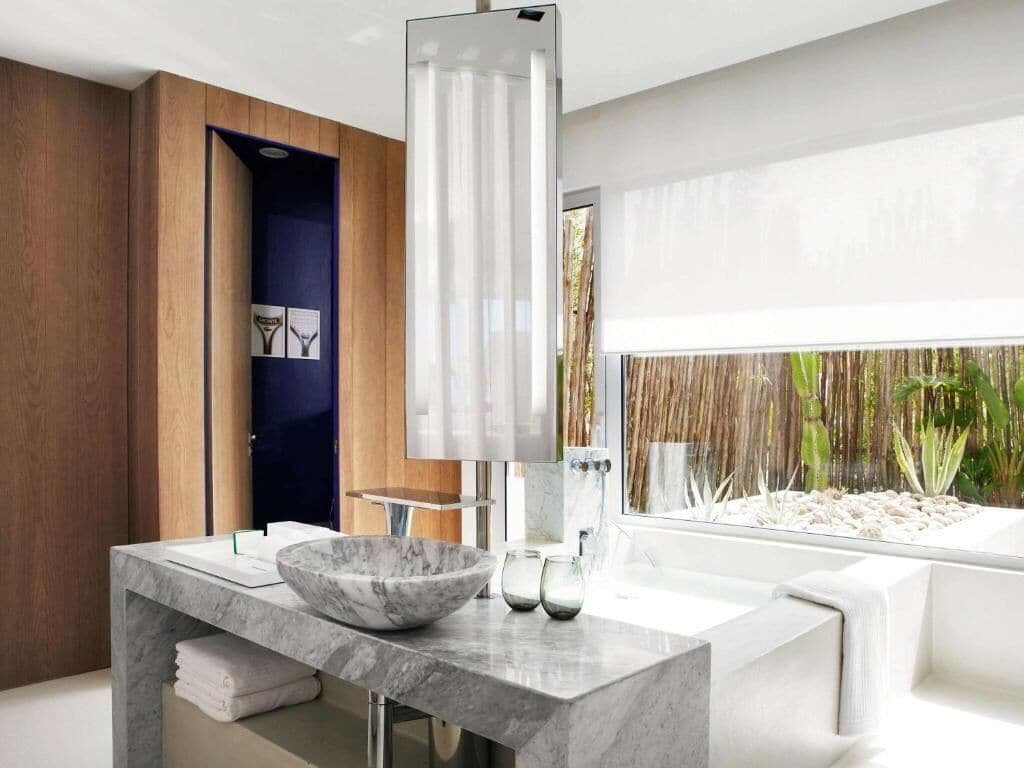 A modern bathroom with a marble sink, a large mirror, and a view of a bamboo fence and plants.