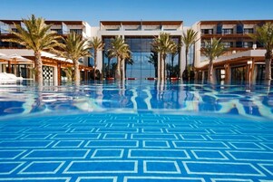 Piscina con un diseño geométrico en el suelo, rodeada de palmeras y un edificio moderno con varios balcones.