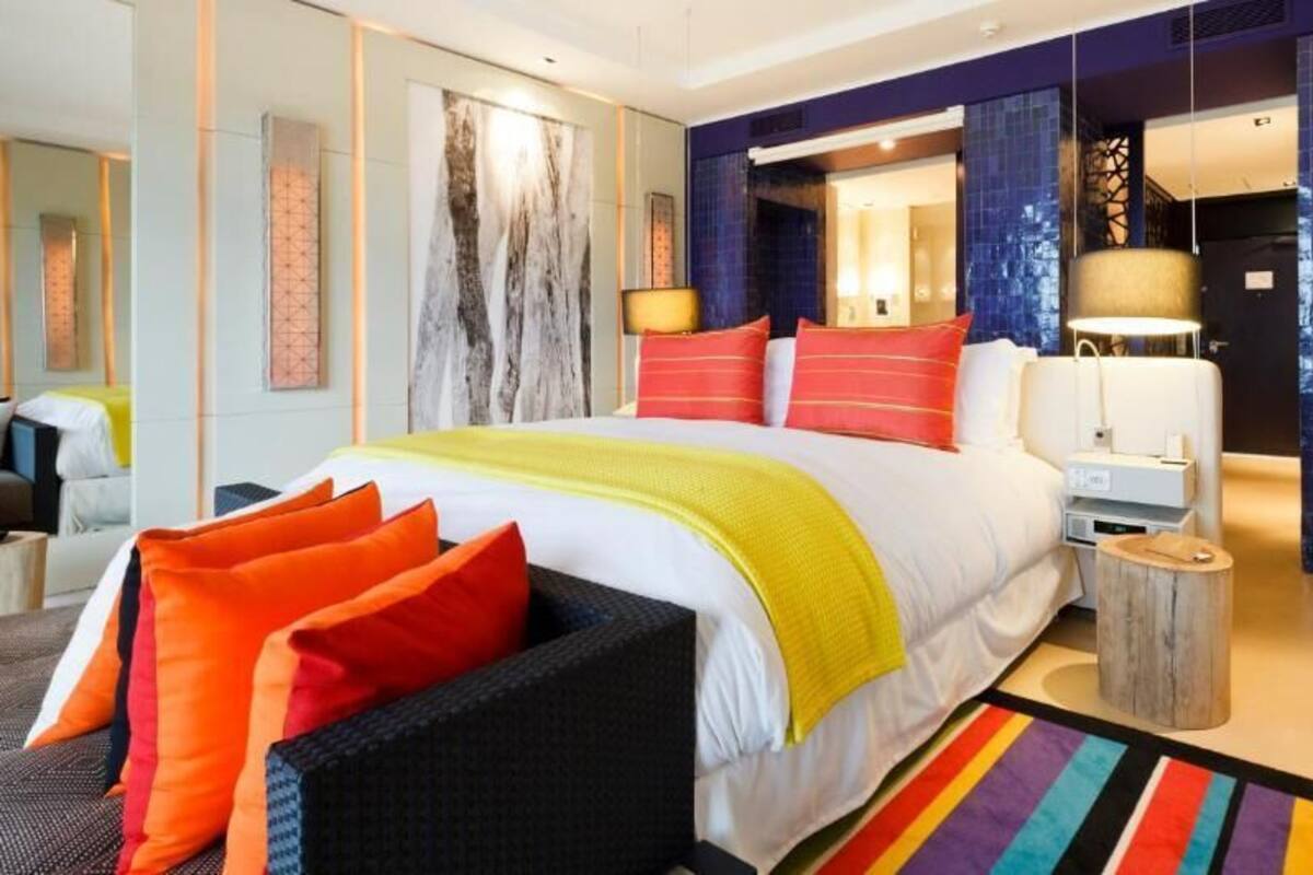 A modern hotel room with a large bed, colorful pillows, a side table, and a vibrant striped rug.