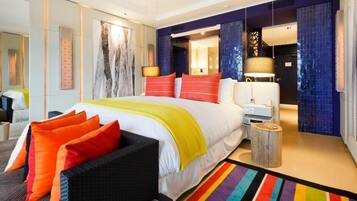 Une chambre d’hôtel moderne avec un grand lit, des oreillers colorés, une table de chevet et un tapis rayé aux couleurs vives.
