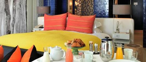 A hotel room with a large bed, breakfast table with fruit and pastries, and a coffee maker.