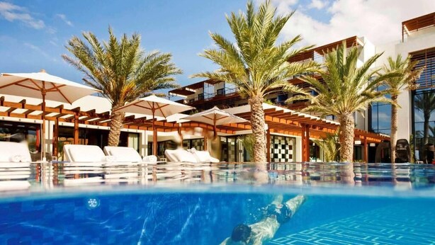 Une piscine entourée de palmiers et de bâtiments modernes, avec des chaises longues et des parasols.