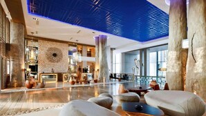 Un hall d’entrée spacieux au design moderne, comprenant un espace de détente central avec des canapés et une table basse, un mur en briques orné d’un motif géométrique et un plafond doté d’un éclairage bleu.