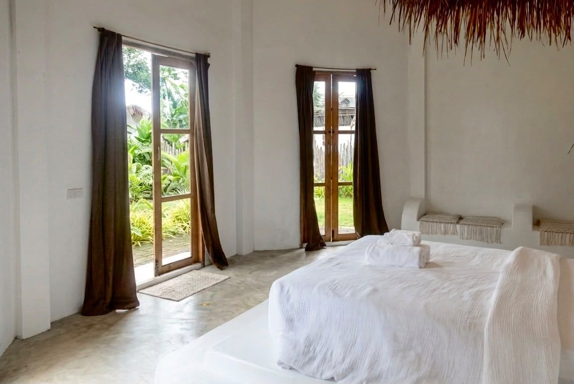 Une chambre avec un lit, deux fenêtres avec des rideaux et une vue sur la verdure à l’extérieur.