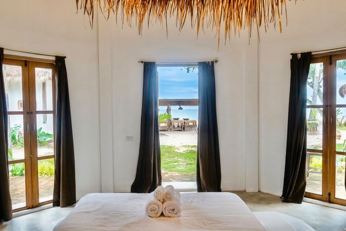 A hotel room with a bed, curtains, and a view of the beach and a wooden bridge.