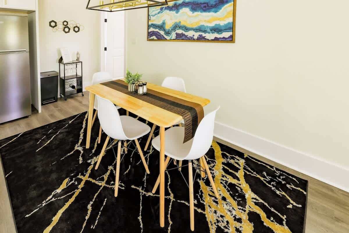 A dining area with a wooden table, white chairs, a black and gold rug, and a painting on the wall.