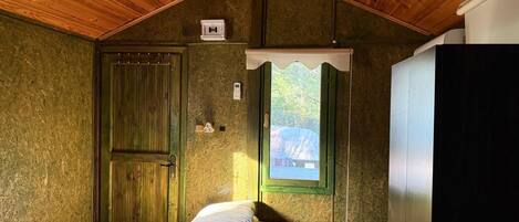A small, cozy room with a wooden ceiling, a bed with white bedding, a green-framed window, and a wardrobe.