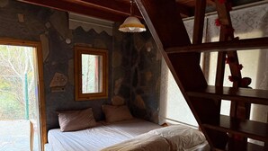 A bedroom with a stone wall, a bed with beige bedding, a window, and a wooden staircase. - Jippe İda Dağ Evleri (Edremit)