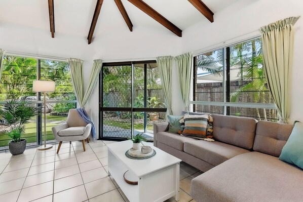 A modern living room with a sofa, armchair, and coffee table, large windows with curtains, and a view of greenery outside.