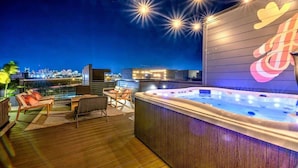 A rooftop terrace with a hot tub, seating area, and a city view at night.