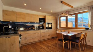 A modern kitchen with wooden cabinets, a dining table with chairs, and a large window with a view of mountains. - Apartment Hipp No. 6 - 77m² new building on the ground floor with terrace and mountain view (Halblech)