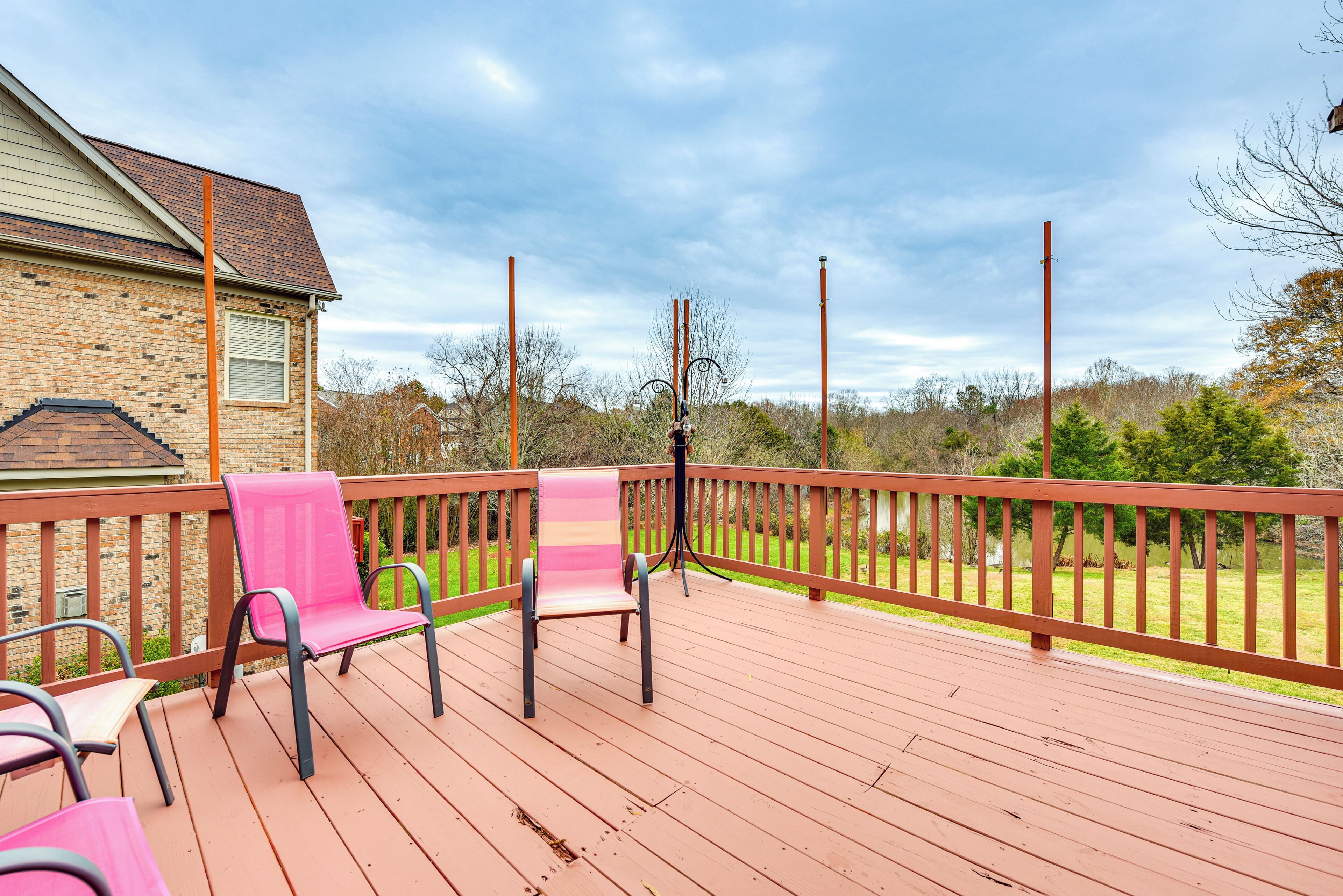 Une terrasse en bois avec deux chaises roses et une table, donnant sur une zone herbeuse avec des arbres et un bâtiment en arrière-plan.
