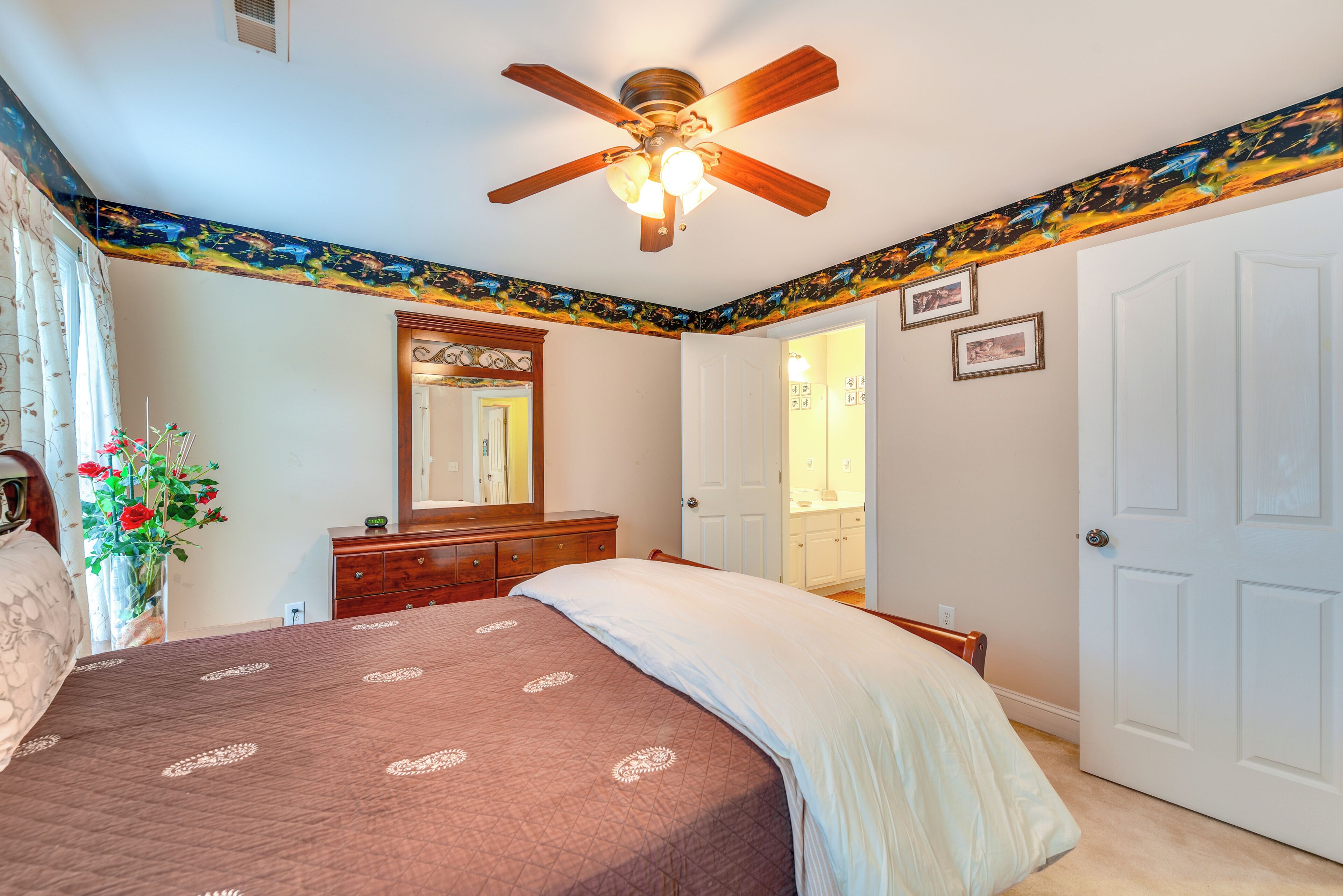 A bedroom with a bed, a dresser, a ceiling fan, a mirror, and a door.