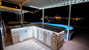 An outdoor kitchen area with a built-in island and a pool in the background. - Pool & Hot Tub Access: Luxury Roy Oasis! (Roy)