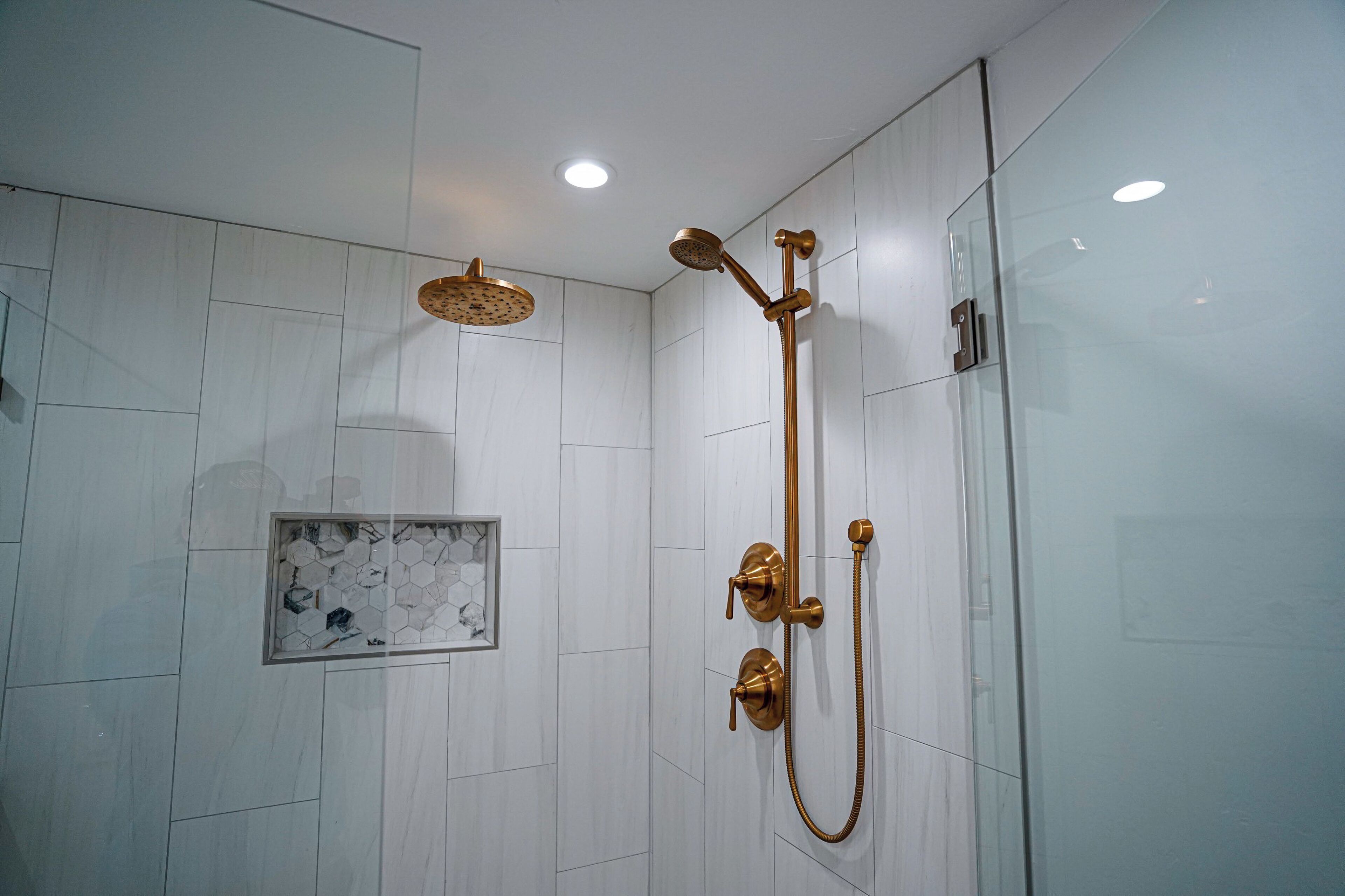 A modern shower with a gold showerhead and handheld showerhead, white tiled walls, and recessed ceiling lights.