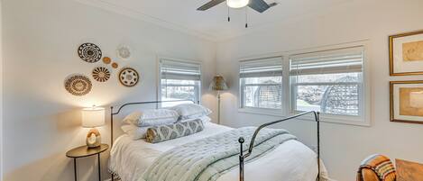 A bedroom with a bed, bedside lamp, ceiling fan, windows, and wall decor.