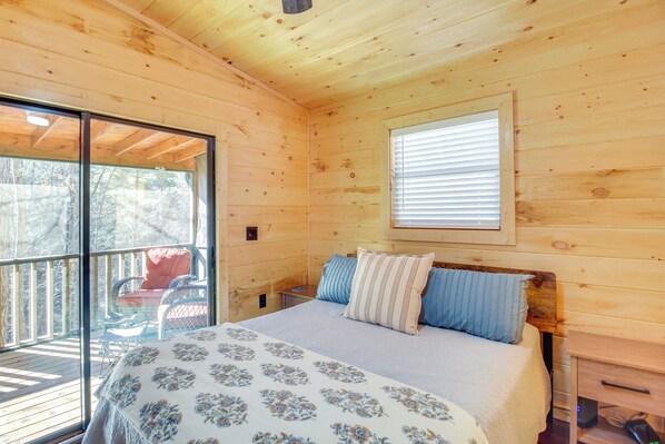 A wooden cabin bedroom with a bed, pillows, a window, and a balcony. - Hot Tub & Creek View at Cabin: 6 Mi to Ellijay! (Ellijay)