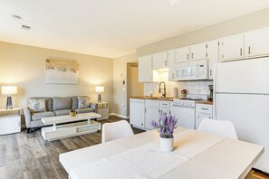 Ein modernes Wohnzimmer mit Sofa, Couchtisch und Esstisch. Die Küche verfügt über weiße Schränke, eine Mikrowelle und einen Kühlschrank.