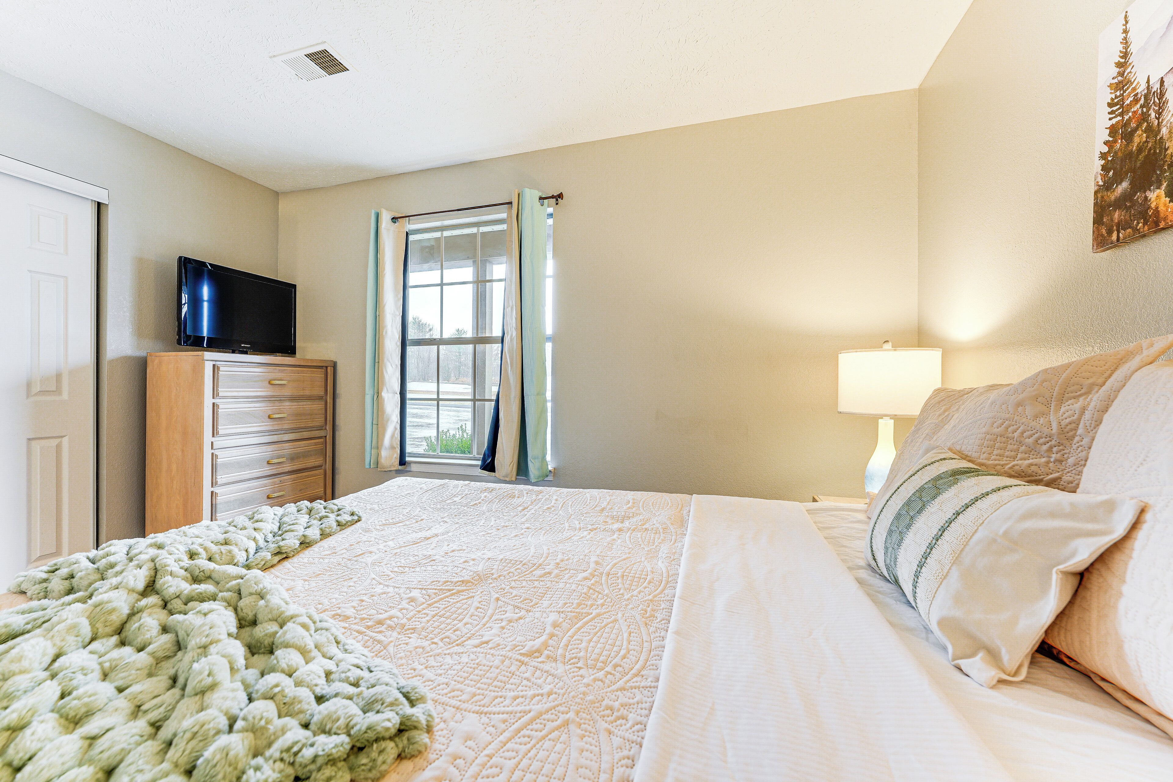 A bedroom with a bed, a dresser, a television on top of the dresser, a window with curtains, and a painting on the wall.