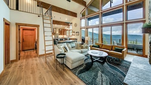 A spacious living area with a wooden floor, a large window offering a mountain view, a staircase, and a kitchen in the background. - Stunning View! Peaceful & Modern Schweitzer Chalet (Sandpoint)