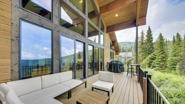 A wooden deck with a sofa and chairs, large windows, and a view of trees and hills. - Stunning View! Peaceful & Modern Schweitzer Chalet (Sandpoint)