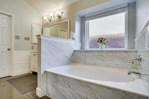 Une salle de bain avec une grande baignoire, une fenêtre à vitres dépolies et un vase rempli de fleurs posé sur le rebord de la fenêtre.