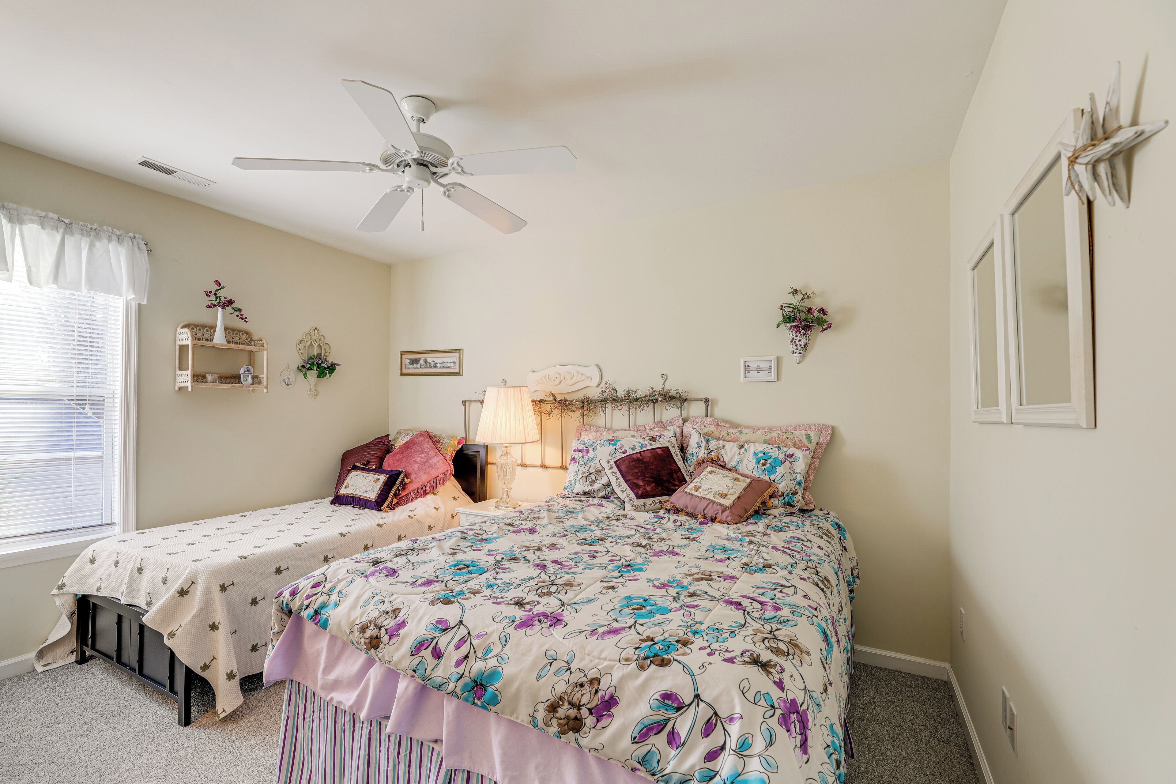A bedroom with two beds, a ceiling fan, a window with blinds, and wall-mounted shelves.