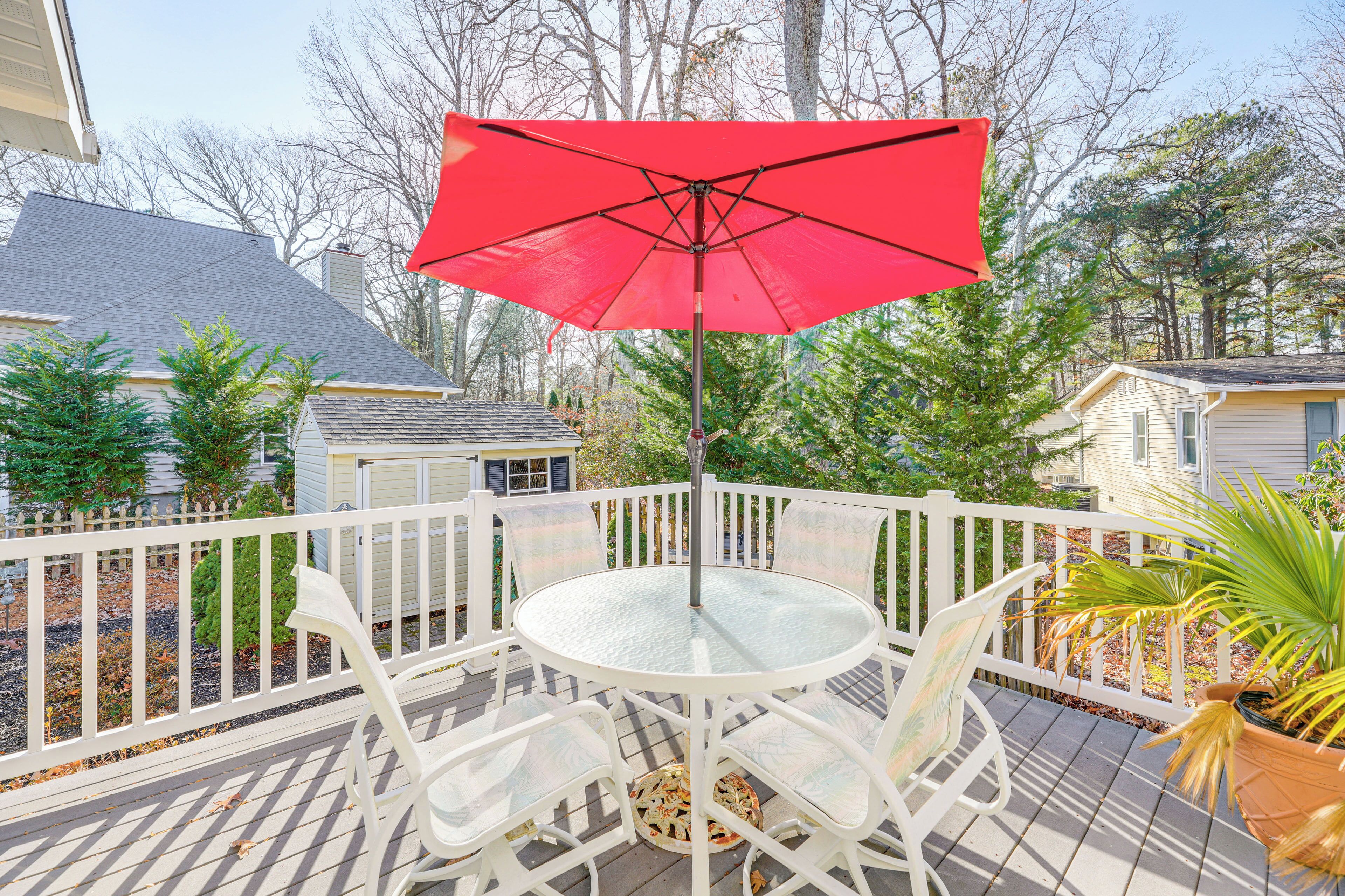 A round table with white chairs and a red umbrella on a wooden deck.