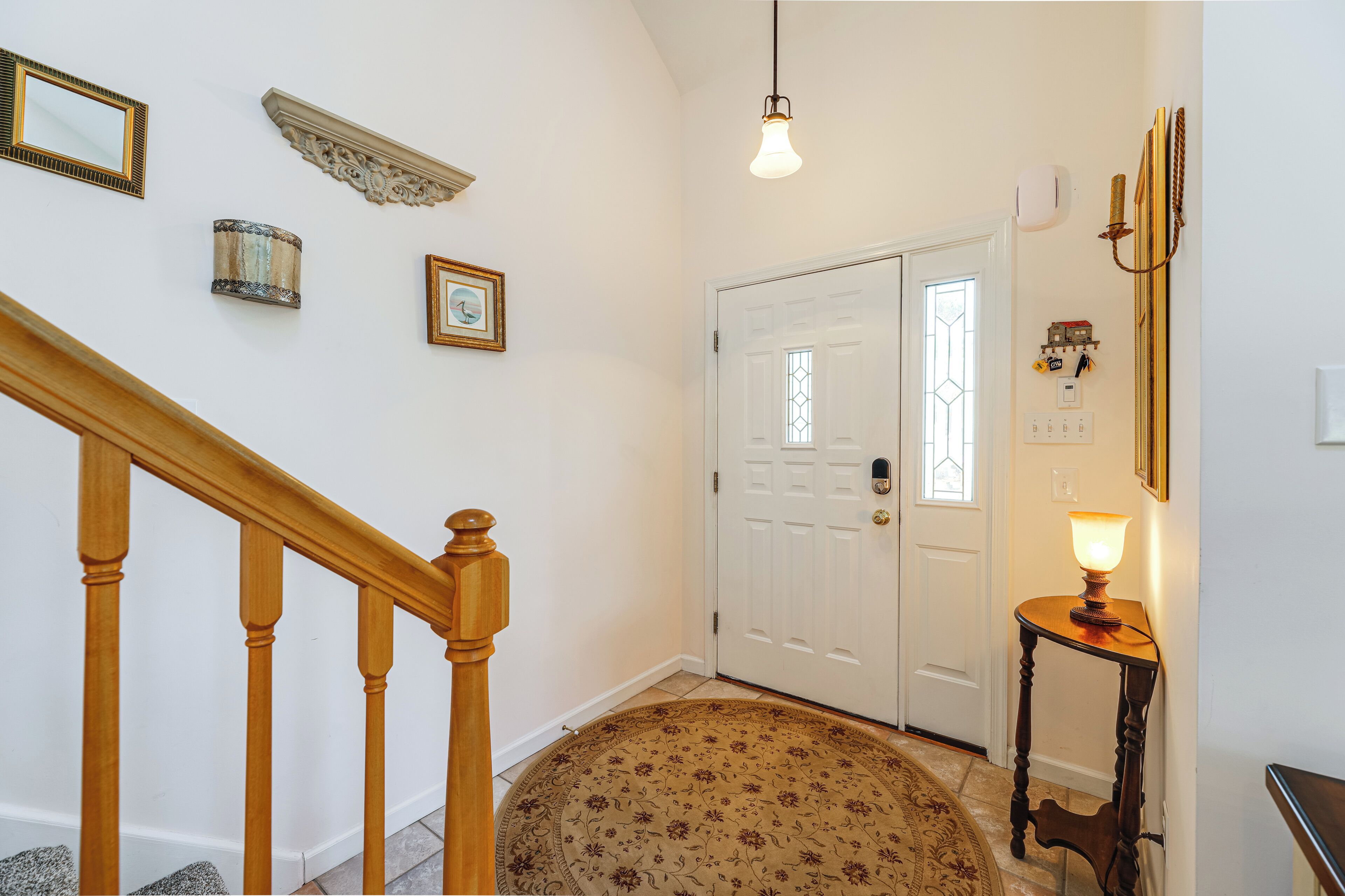A hallway with a wooden staircase, a white door with a glass window, a round carpet, and a small table with a lamp.