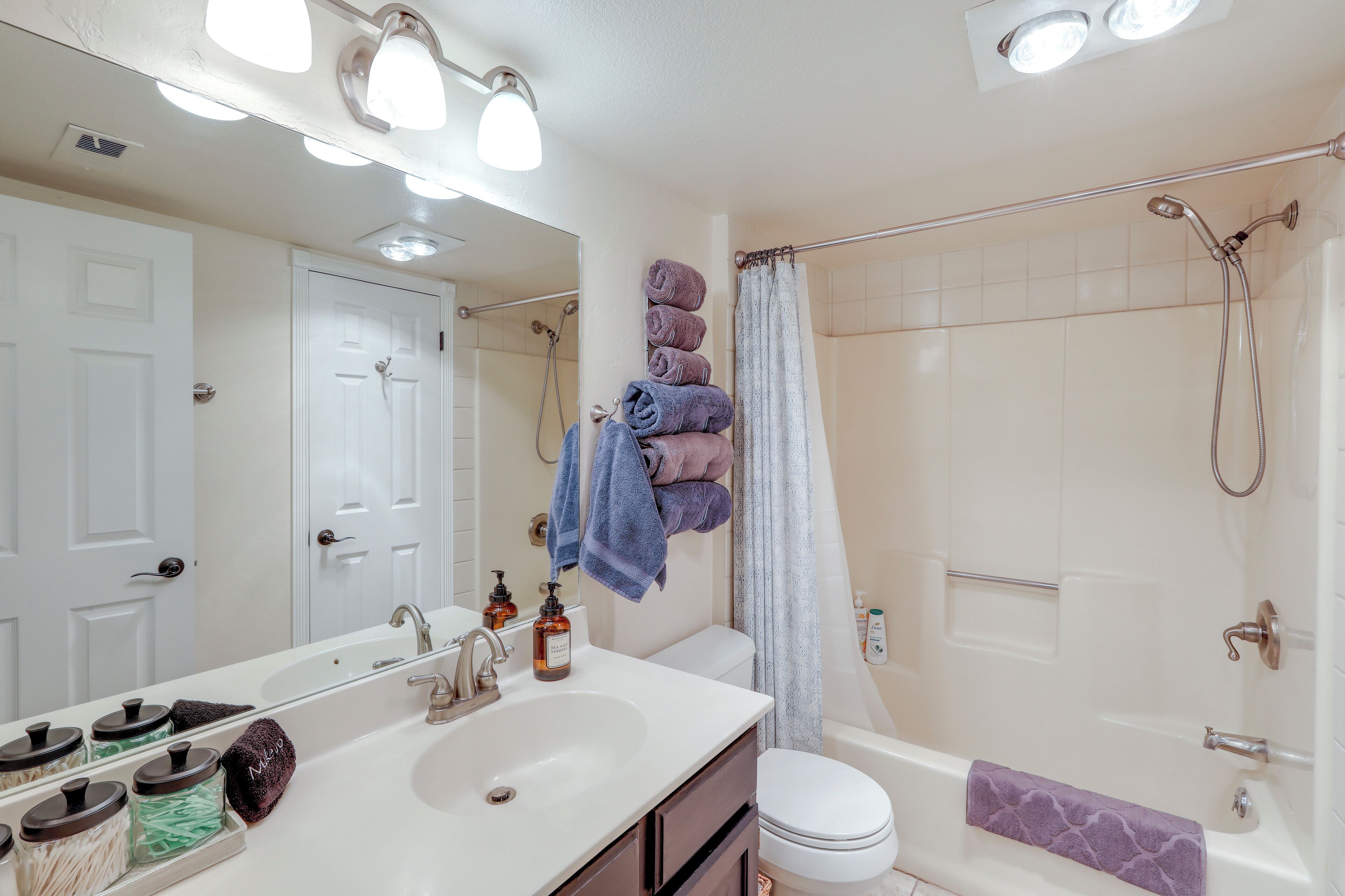 Une salle de bain avec un lavabo, un miroir et une douche. Des serviettes sont pliées et empilées sur un porte-serviettes au mur, et il y a un rideau de douche.